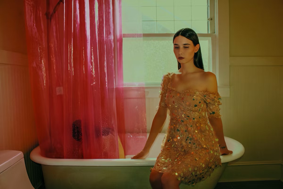 Sad woman sitting on the edge of a vintage bathtub, dressed in a sparkling, multi-coloured dress, with a pink shower curtain in the background.
