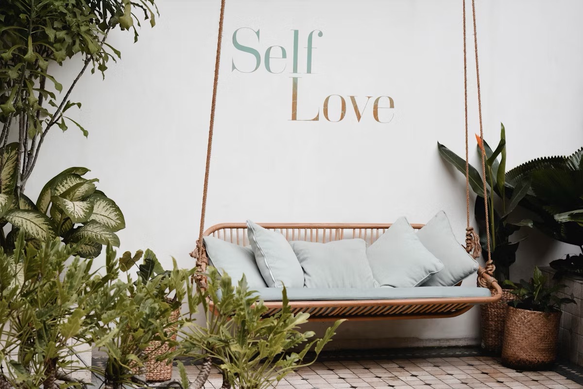 A cosy swing with soft blue cushions surrounded by greenery, featuring the inspirational text "Self Love" on a white wall, symbolising the theme of embracing authenticity.
